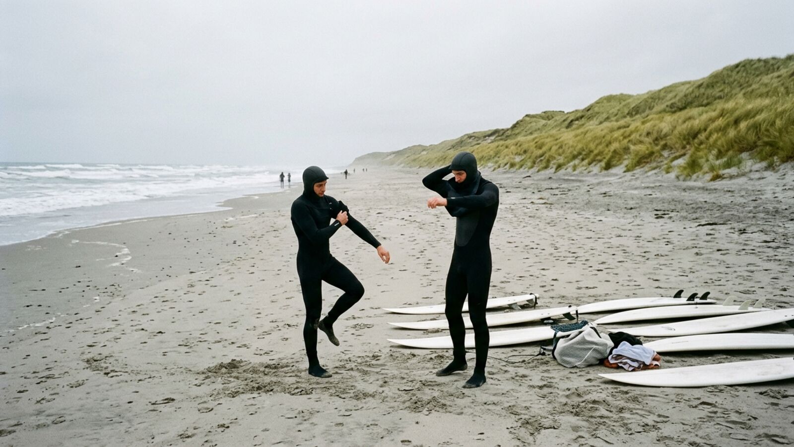 Deux surfeurs enfilent leur combinaison intégrale sur une plage de dunes, planches posées sur le sable